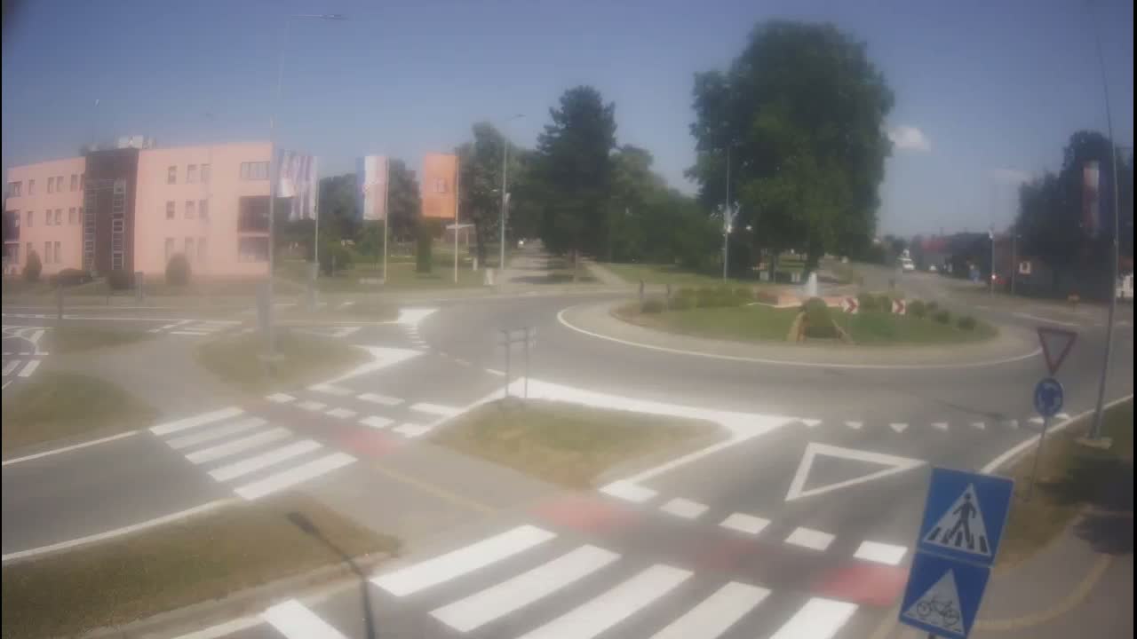 A roundabout intersection with crosswalks and pedestrian and bicycle signage is shown on a sunny day, near a light-pink building.