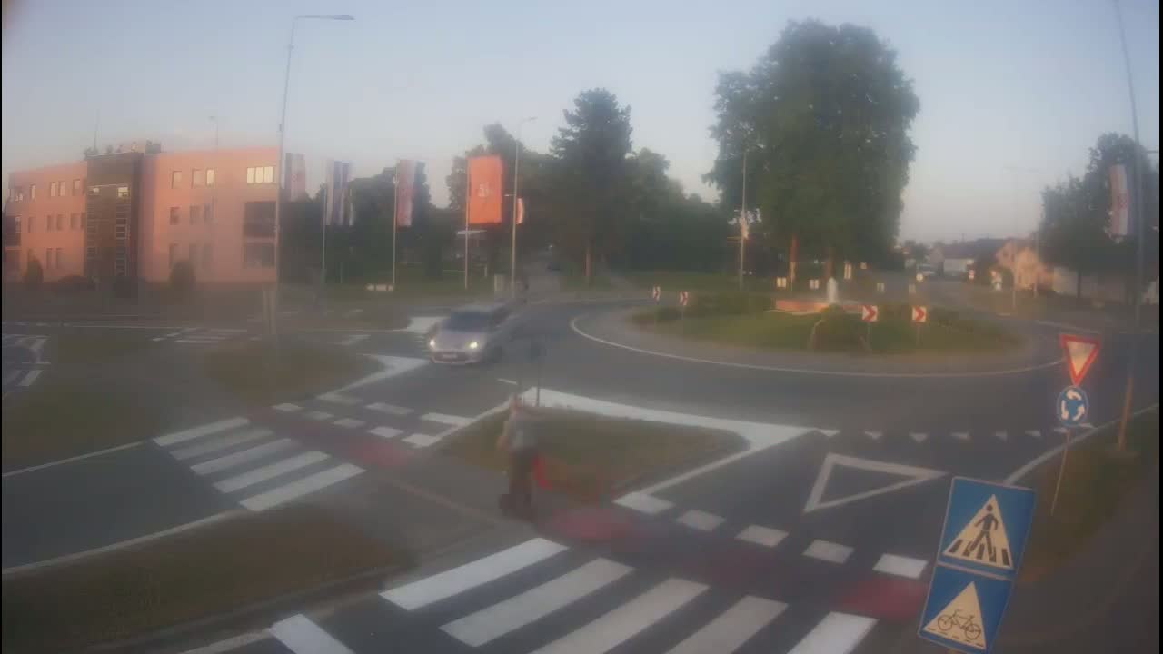 A hazy morning scene shows a roundabout with a car and pedestrian, near a building and some trees.