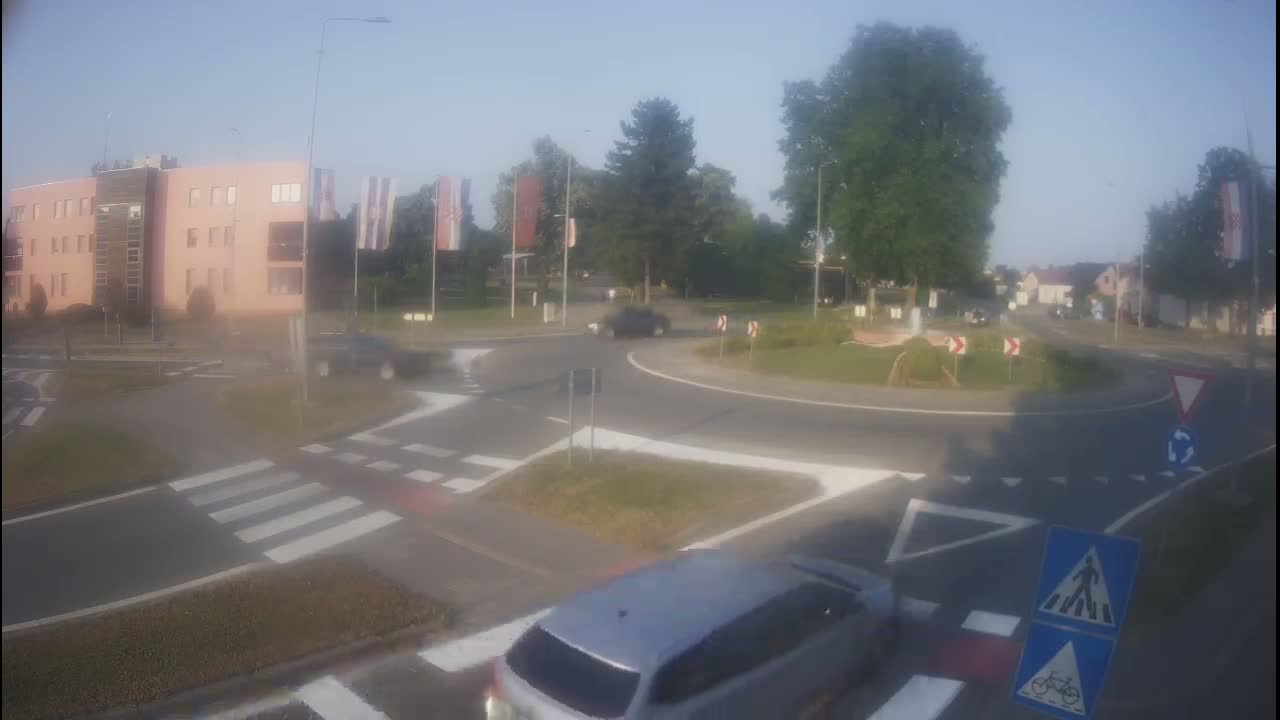 A hazy, sunny day shows a roundabout with several cars, a pedestrian crossing, and a building in the background.