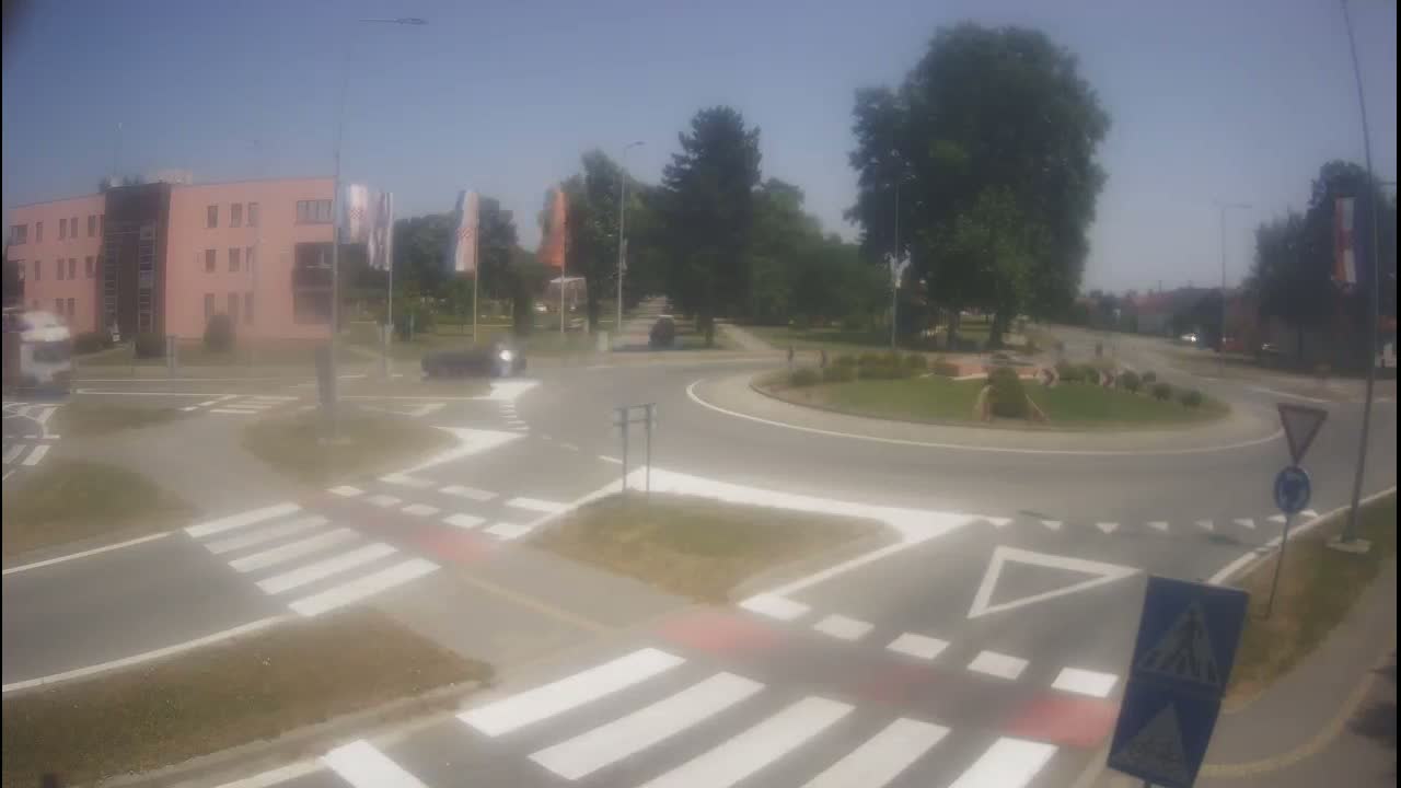 A roundabout with crosswalks and a few cars is seen on a sunny day, surrounded by buildings and trees.