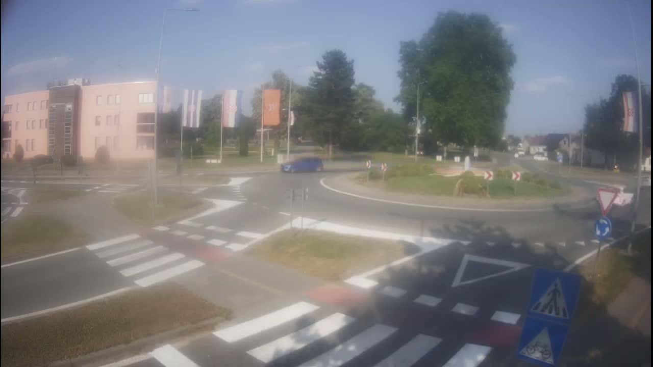 A blue car drives through a roundabout near a pink building on a sunny day.