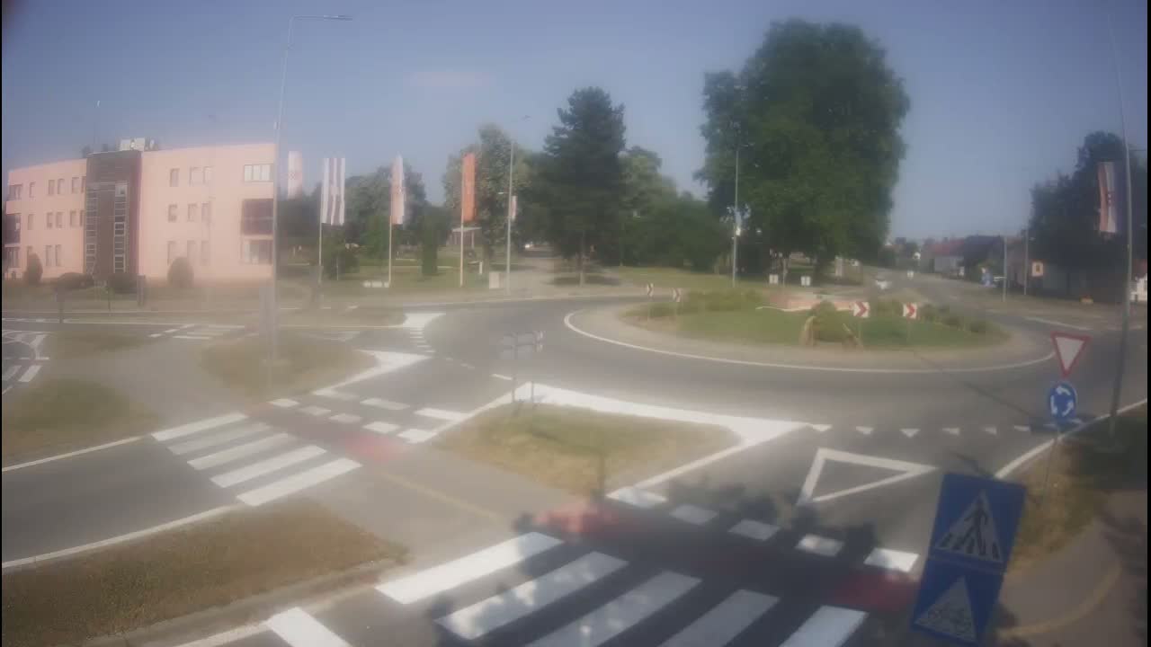 A roundabout with pedestrian crossings is situated near a light pink building under a clear, sunny sky.
