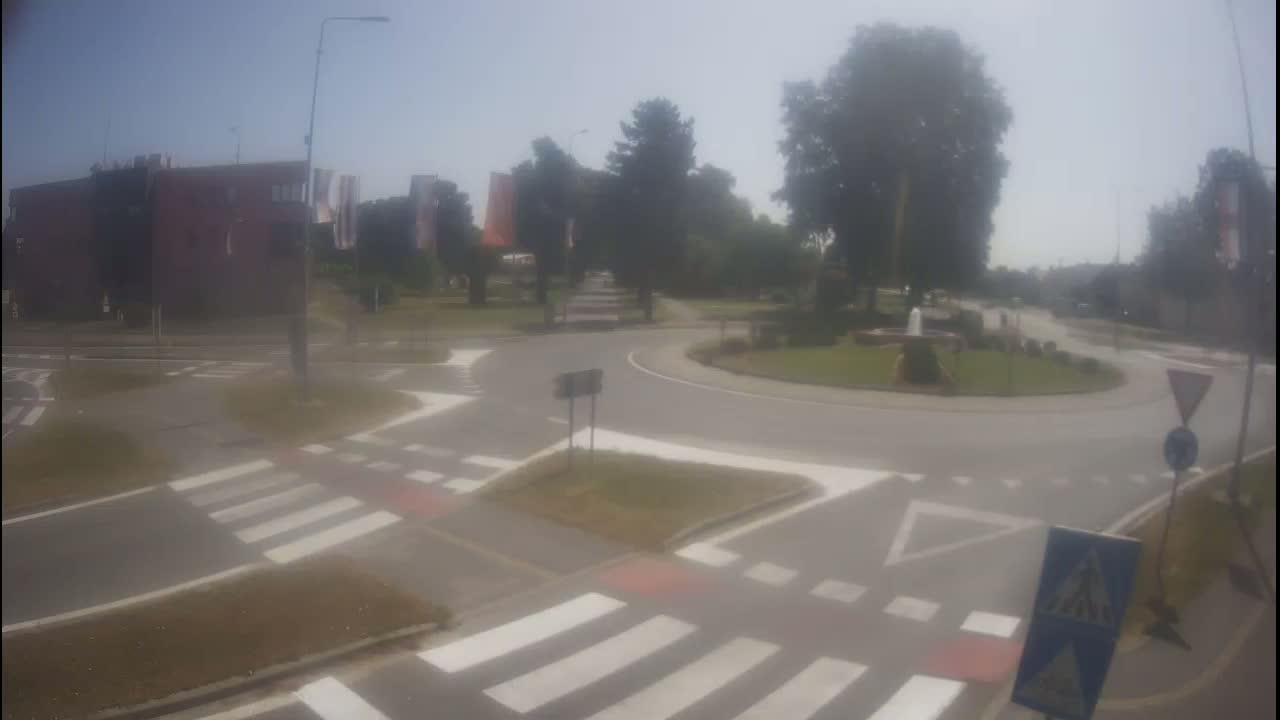 A hazy, sunny day reveals a roundabout intersection with crosswalks and a small fountain in the center, surrounded by trees and buildings.