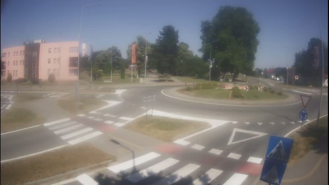 A roundabout with pedestrian crossings is shown on a sunny day, next to a light-pink multistory building and a green area with trees.