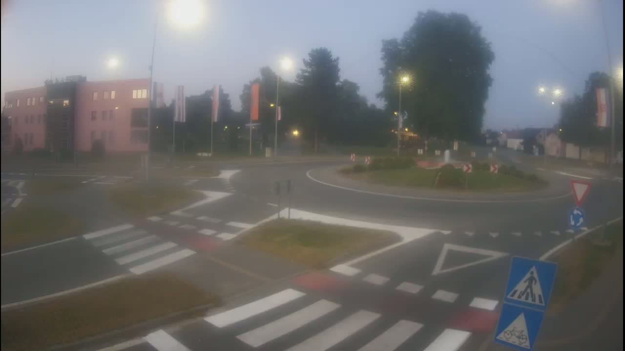 A roundabout with pedestrian crossings is shown on a sunny day, next to a light-pink multistory building and a green area with trees.