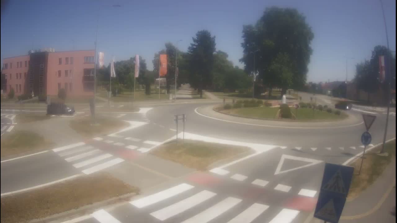 A roundabout with pedestrian crossings is situated near a pink building under a sunny sky.