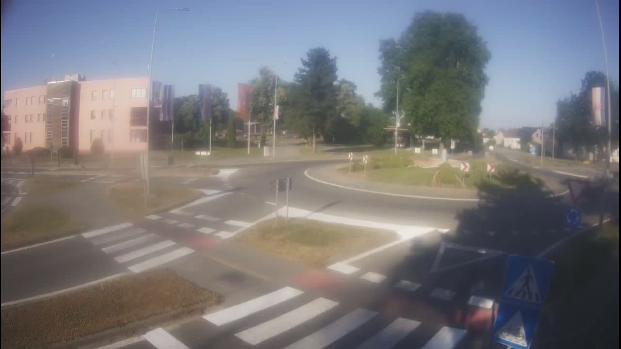 A roundabout intersection on a sunny day, with a light-pink building visible in the background.