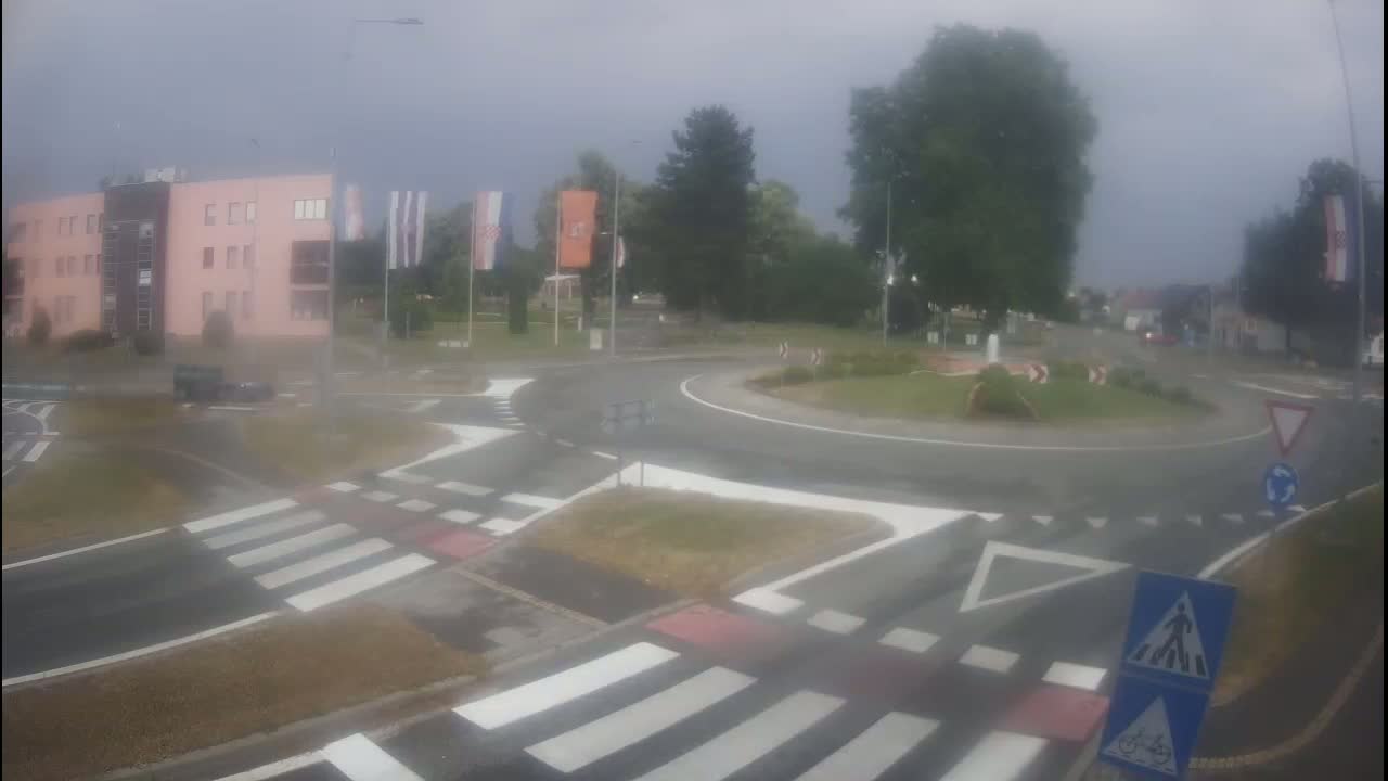 A wet roundabout with pedestrian crossings is seen on an overcast day, next to a pink building and some trees.
