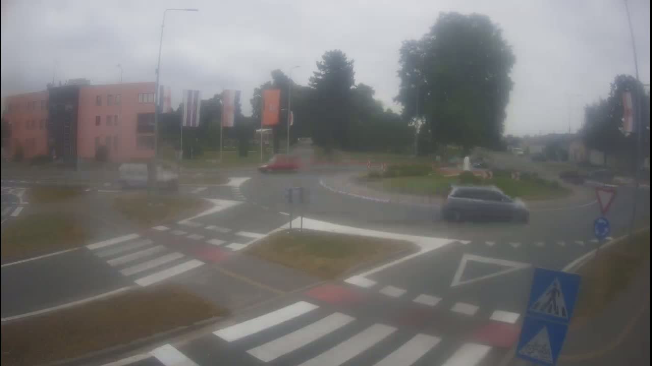 A rainy day scene shows several cars navigating a roundabout near a pedestrian crossing and a light pink building.