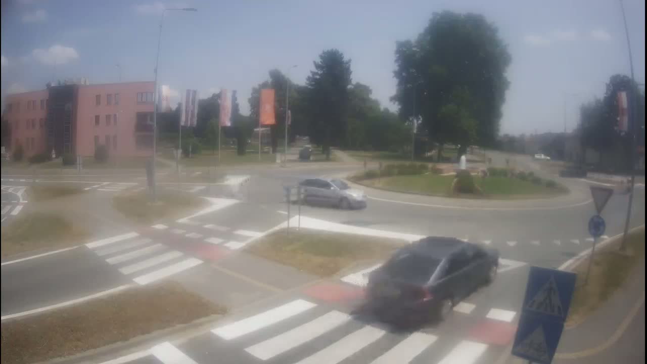 Two cars navigate a roundabout on a sunny day, with a pink building and green trees visible in the background.