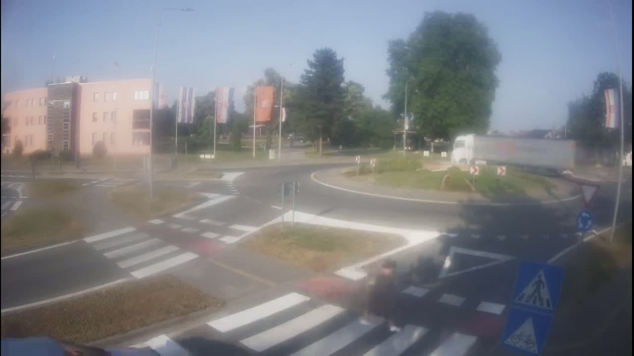 A semi-truck circles a roundabout near a light-pink building on a hazy, sunny day.