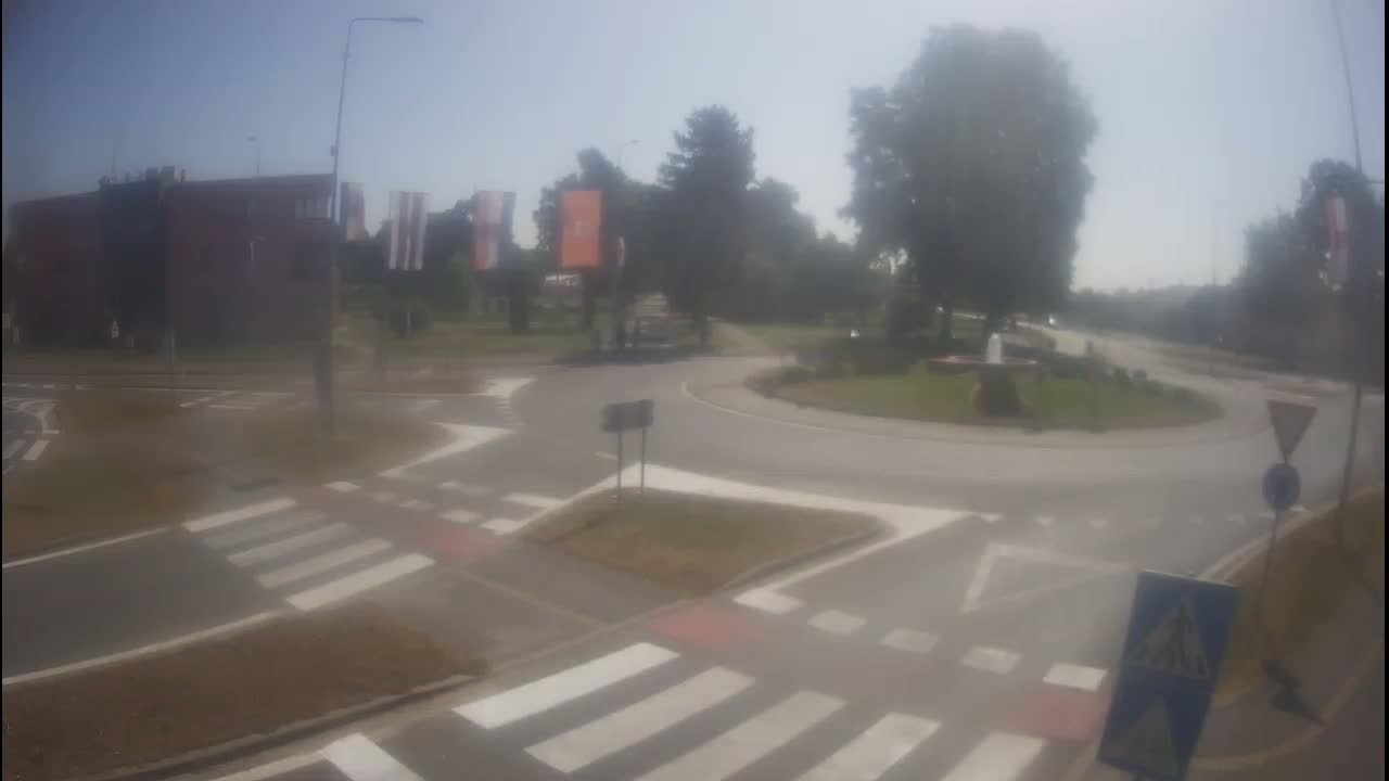 A hazy, sunny day shows a roundabout intersection with crosswalks, a building, and trees.