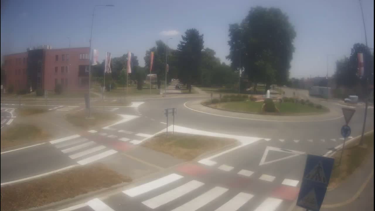 A roundabout with pedestrian crossings is surrounded by a building, trees, and a sunny sky.