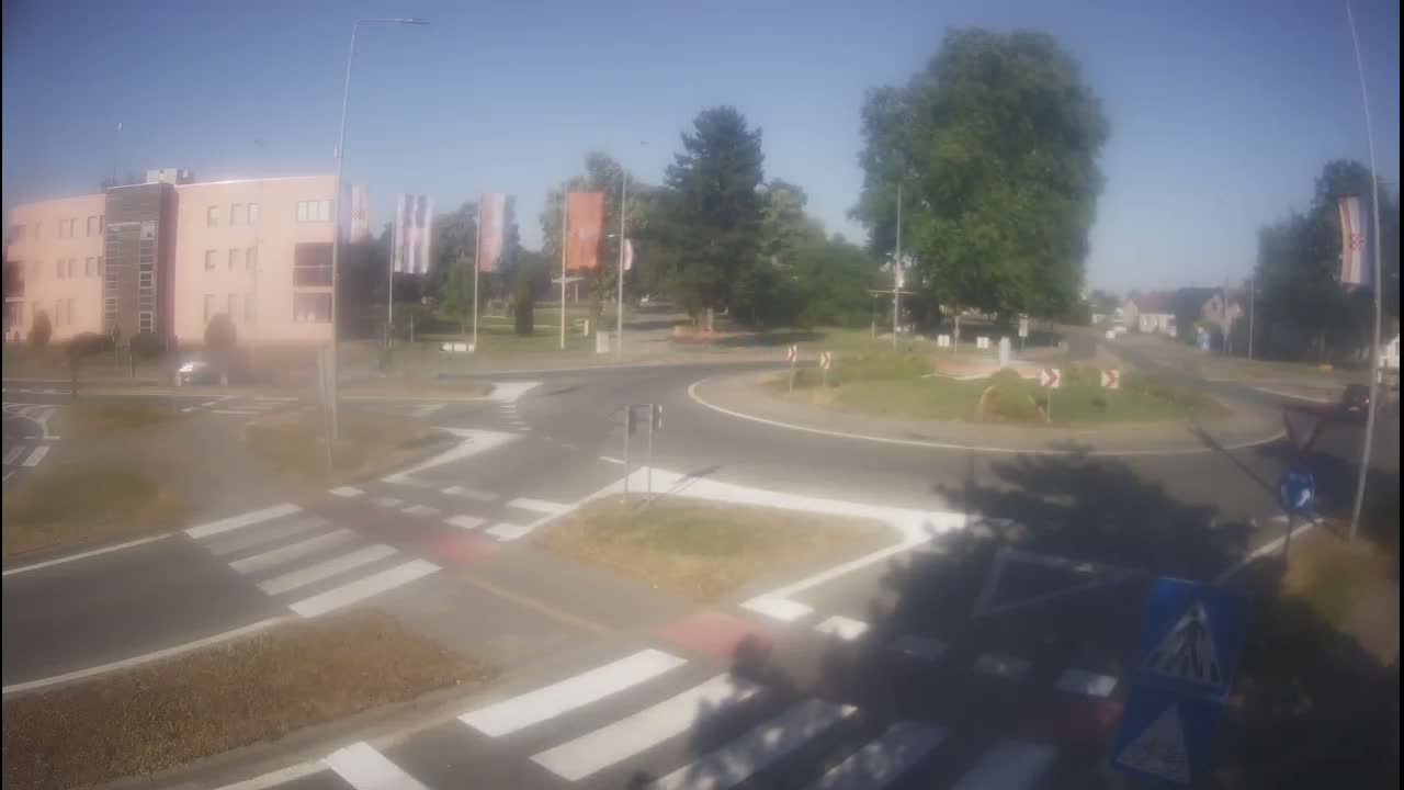 A roundabout intersection on a sunny day, featuring a pink building, pedestrian crossings, and several trees.