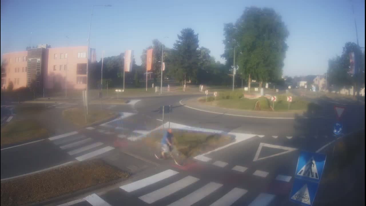 A hazy, sunny day reveals a roundabout with a cyclist crossing a pedestrian crossing near a light-pink building.