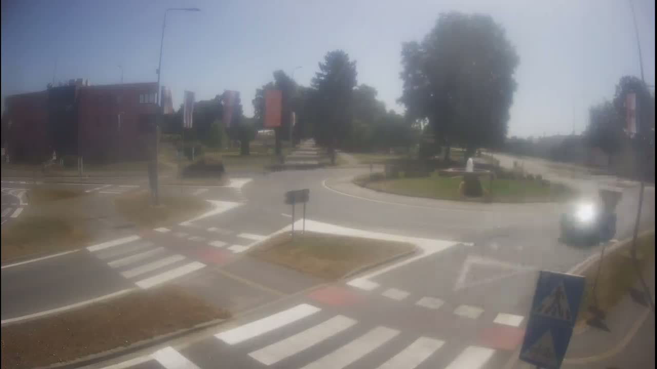 A hazy, sunny day reveals a roundabout intersection with pedestrian crossings and a small fountain in the center, surrounded by buildings and greenery.