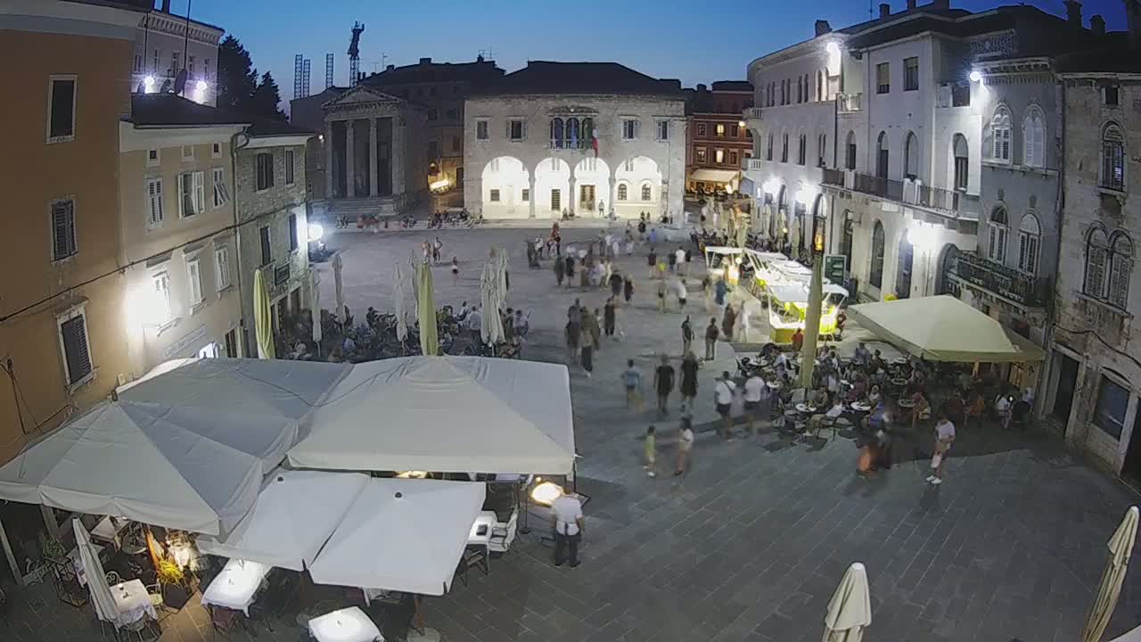 A town square at night, illuminated by lights, is filled with people walking and sitting at outdoor cafes under market umbrellas.