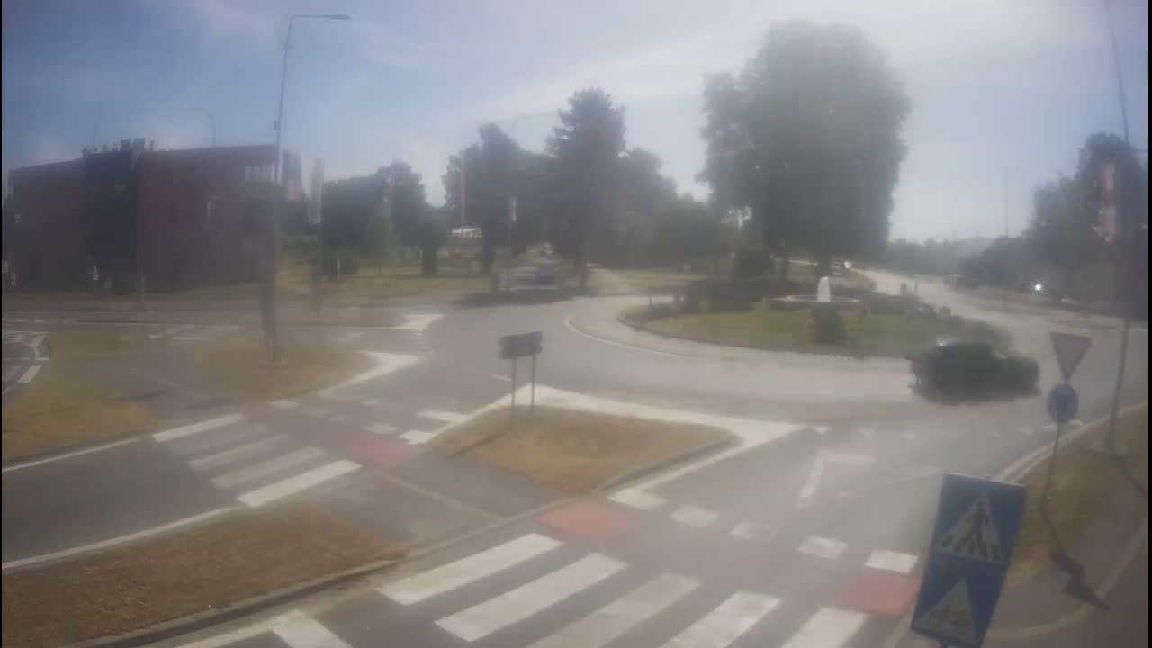 A hazy, sunny day reveals a roundabout intersection with pedestrian crossings, a few cars, and surrounding greenery including trees and a building.