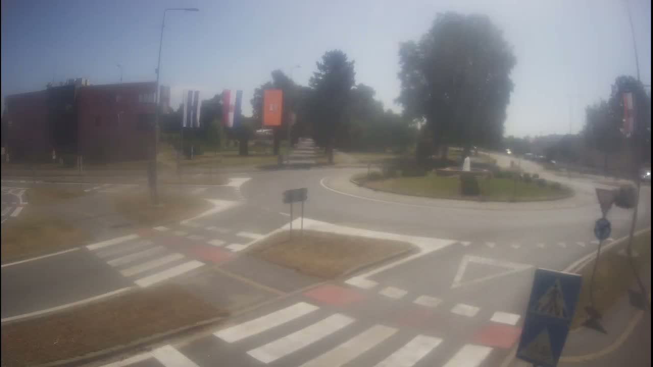 A hazy, sunny day reveals a roundabout intersection with pedestrian crosswalks, a building, and greenery.