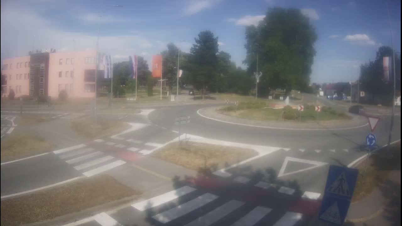 A hazy, sunny day reveals a roundabout intersection with pedestrian crosswalks, a building, and greenery.