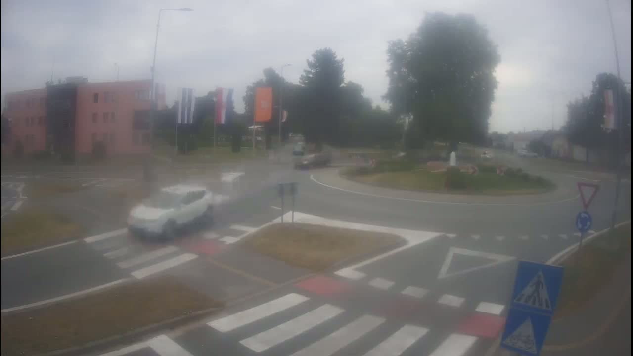 A white car drives through a roundabout on a cloudy day, with other vehicles visible in the distance.
