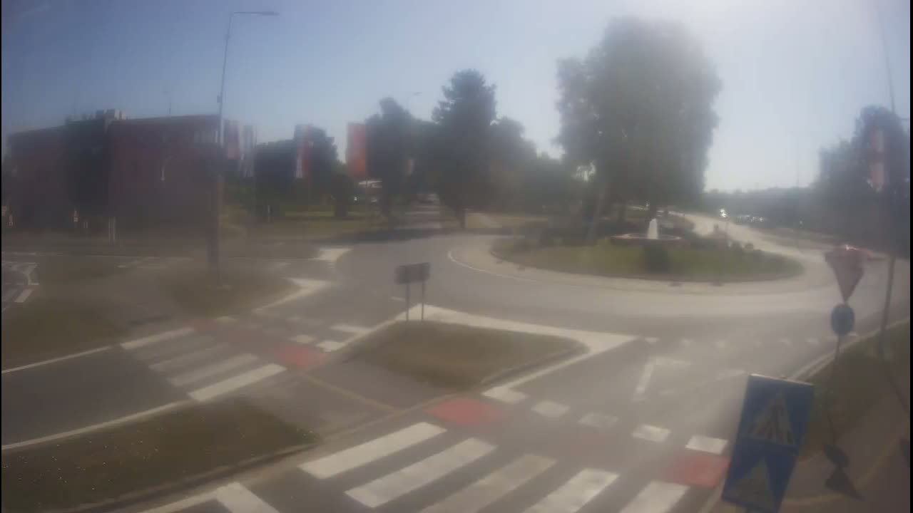 A hazy sunny day shows a roundabout with pedestrian crossings and a small fountain in the center, surrounded by buildings and trees.