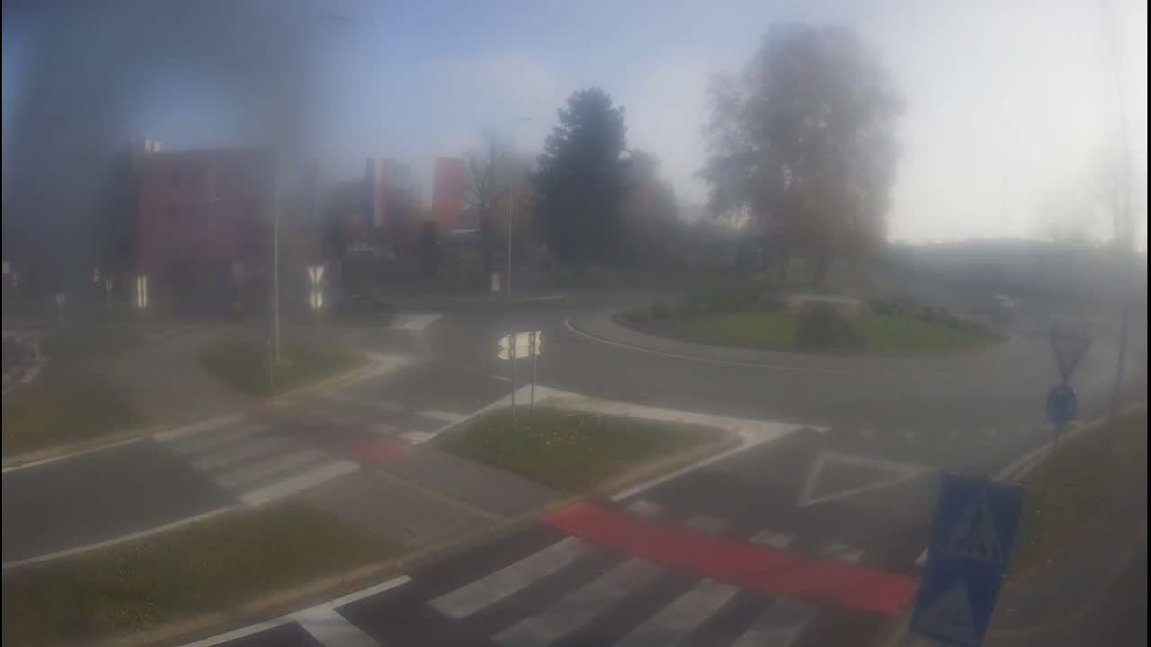 A hazy and blurry outdoor scene shows a quiet urban intersection with a central roundabout, roads, crosswalks, buildings, and trees, all obscured by dense fog.