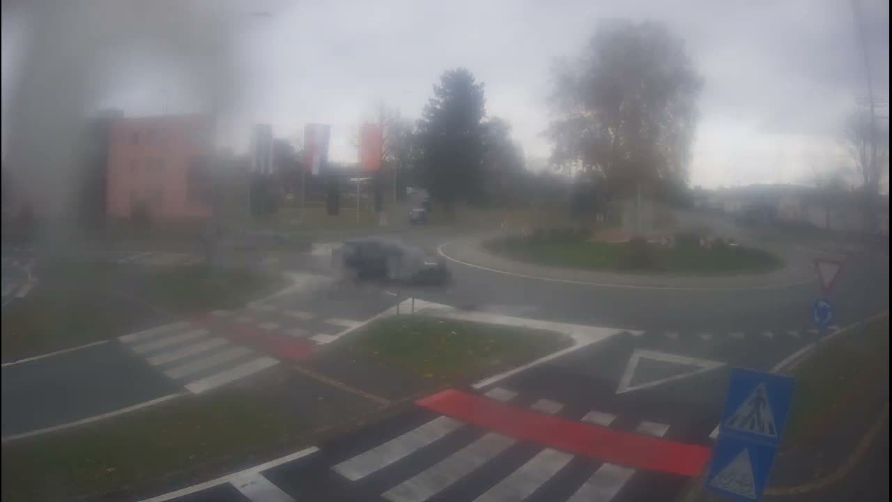 A wet and overcast outdoor scene depicts a car traveling through a roundabout marked with pedestrian crossings, alongside blurred buildings and trees in the background.