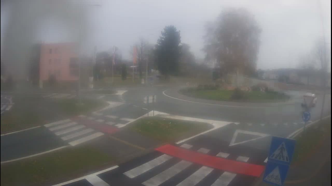 I see a hazy, outdoor view of a roundabout intersection with numerous pedestrian crossings and surrounding greenery, a distant building, and trees, all under very overcast and wet conditions, likely due to rain or mist on the camera lens.