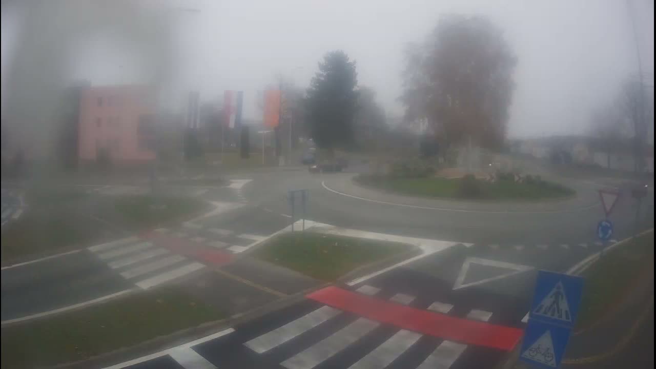 Thick fog envelops a road intersection featuring a roundabout with a dark car driving through it, alongside prominent pedestrian crossings, scattered trees, and indistinct buildings in the background.