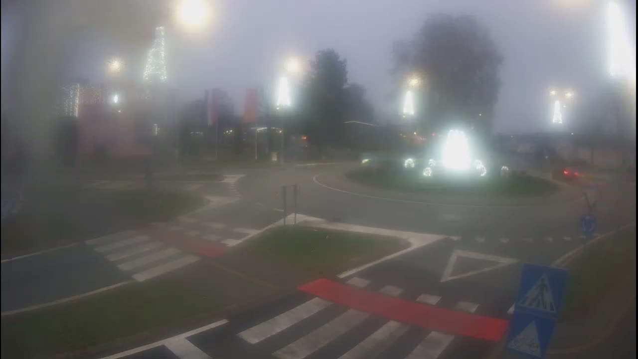 Thick fog envelops a road intersection featuring a roundabout with a dark car driving through it, alongside prominent pedestrian crossings, scattered trees, and indistinct buildings in the background.