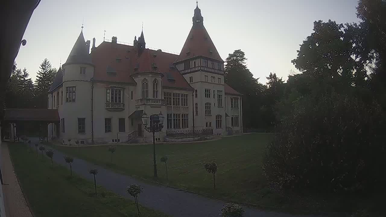 A large, light-colored castle with a red roof sits on a grassy lawn surrounded by trees under a dusky sky.