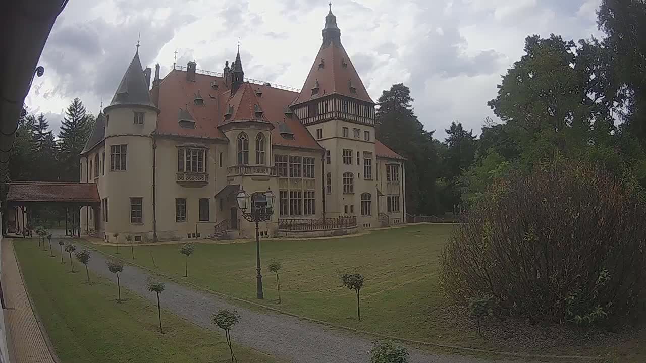 A large, light-colored castle with a red tile roof stands on a grassy lawn under a cloudy sky.