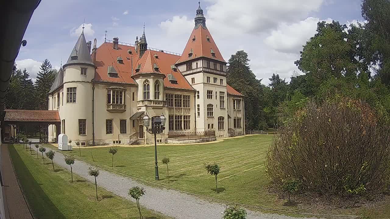 A large, light-colored castle with a red tile roof stands on a grassy lawn under a partly cloudy sky.