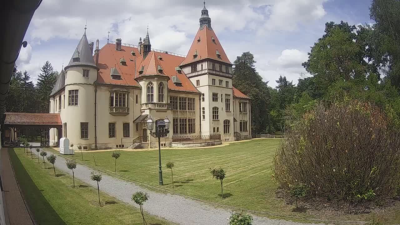 A large, light-colored castle with a red roof sits on a grassy lawn on a partly cloudy day.