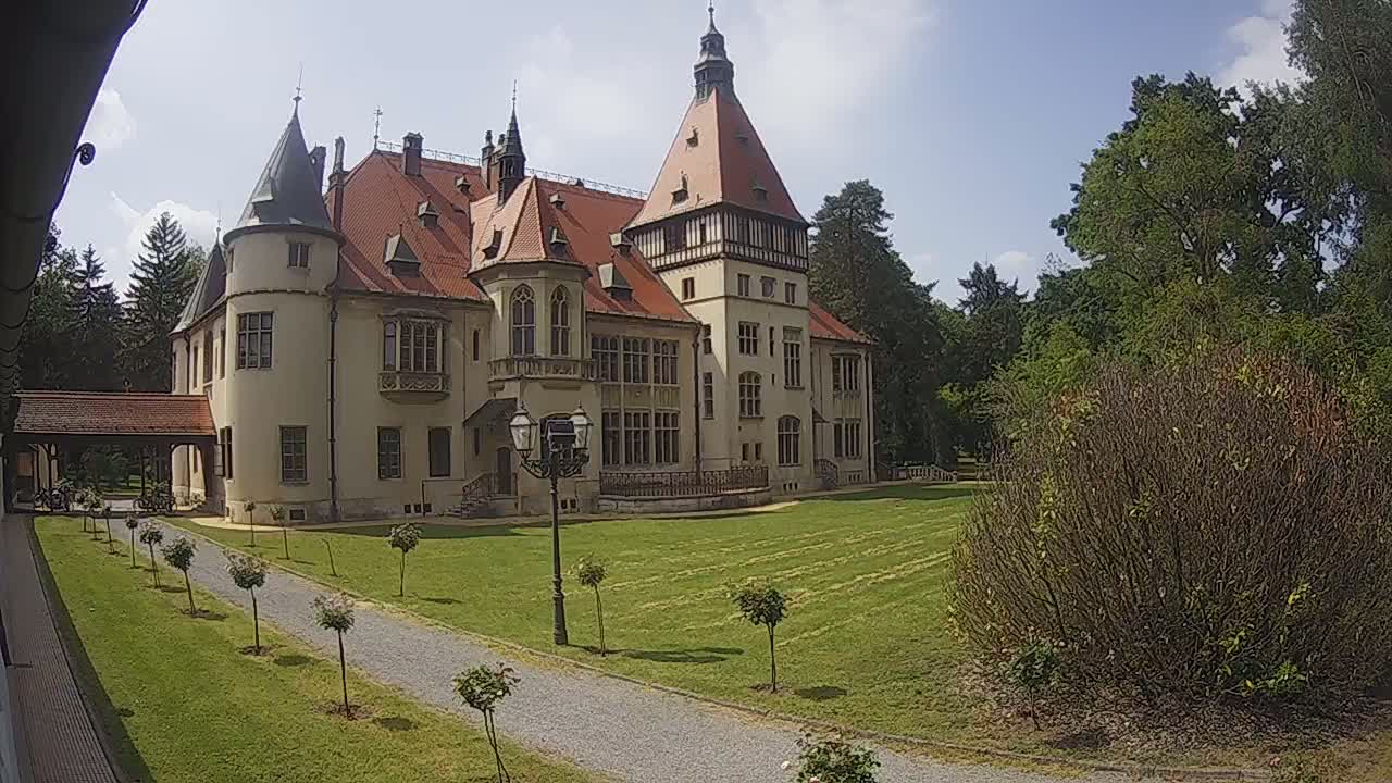 A large, light-colored castle with a red-tiled roof stands on a grassy lawn under a mostly sunny sky, with a gravel path leading to it from the foreground.