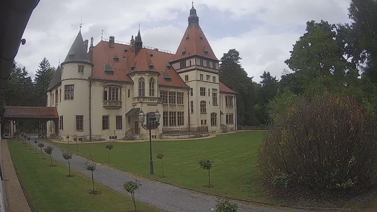 A large, light-colored castle with a red tile roof sits on a grassy lawn under an overcast sky.