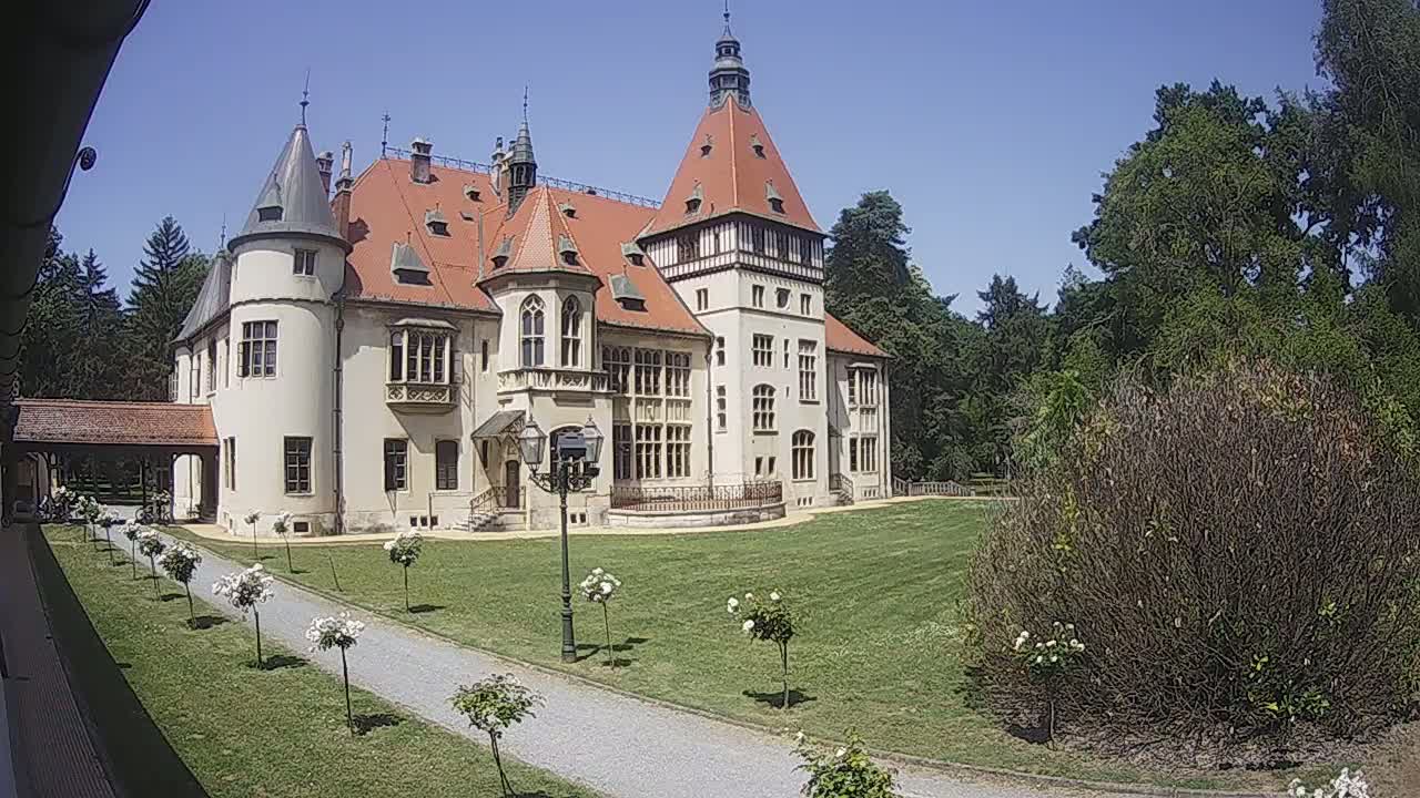 A large, light-colored castle with a red tile roof and multiple towers sits on a grassy lawn on a sunny day, surrounded by trees and neatly planted shrubs.