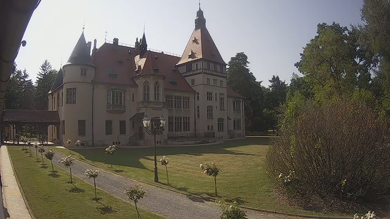 A large, light-colored castle with a red roof stands on a grassy lawn on a sunny day, surrounded by trees and bushes, with a gravel path leading towards it.
