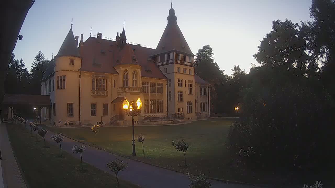 A large, light-colored castle with a red-tiled roof stands on a grassy lawn on a sunny day, surrounded by trees and bushes.