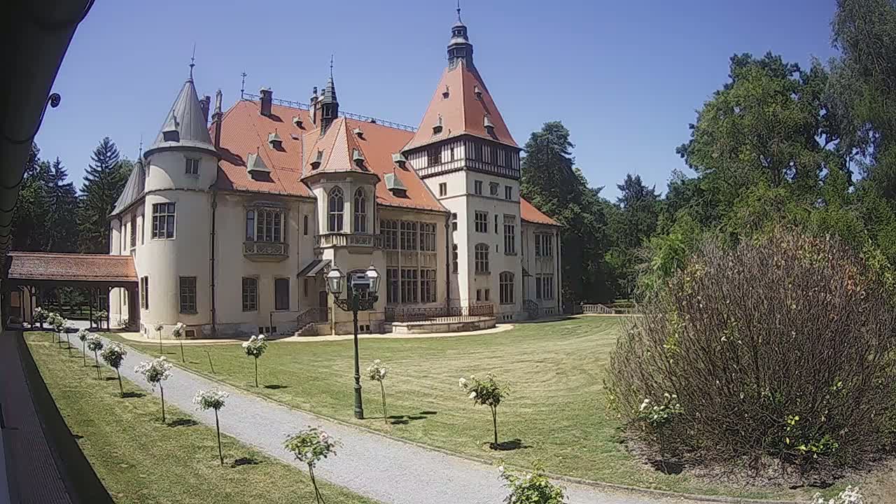 A large, light-colored castle with a red-tiled roof sits on a grassy lawn on a sunny day, with a gravel path leading towards it and several small trees and bushes lining the path.