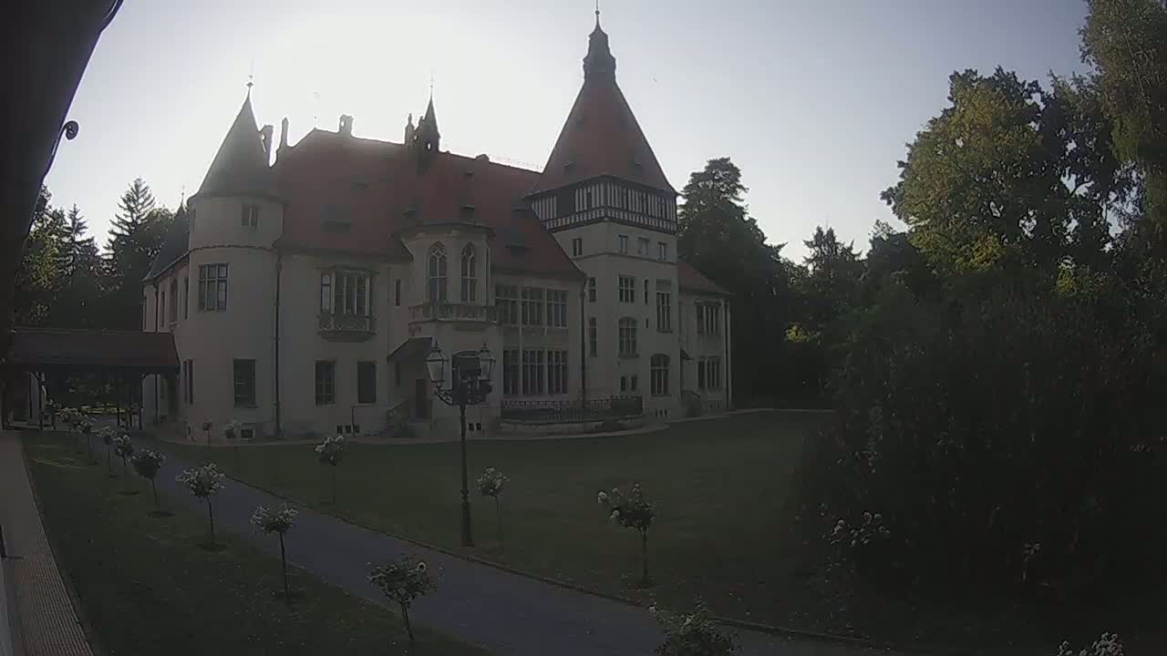 A large, light-colored castle-like building with a red roof stands on a grassy lawn surrounded by trees under a bright, clear sky.
