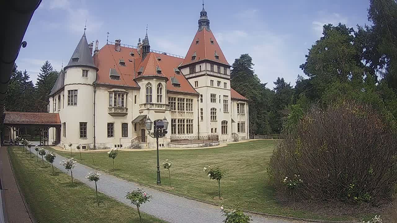 A large, light-colored castle with a red-tiled roof sits on a grassy lawn under a mostly clear sky.