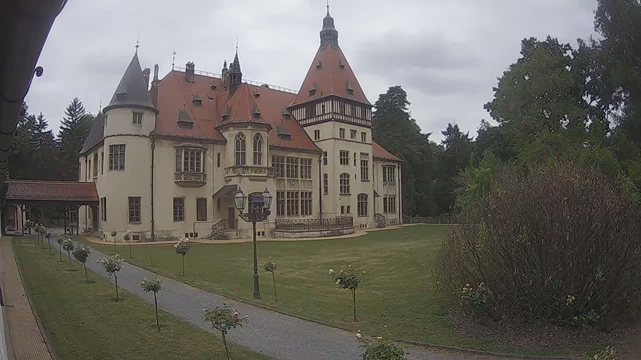 A large, light-colored castle with a red roof stands on a grassy lawn under an overcast sky, surrounded by trees and shrubs.