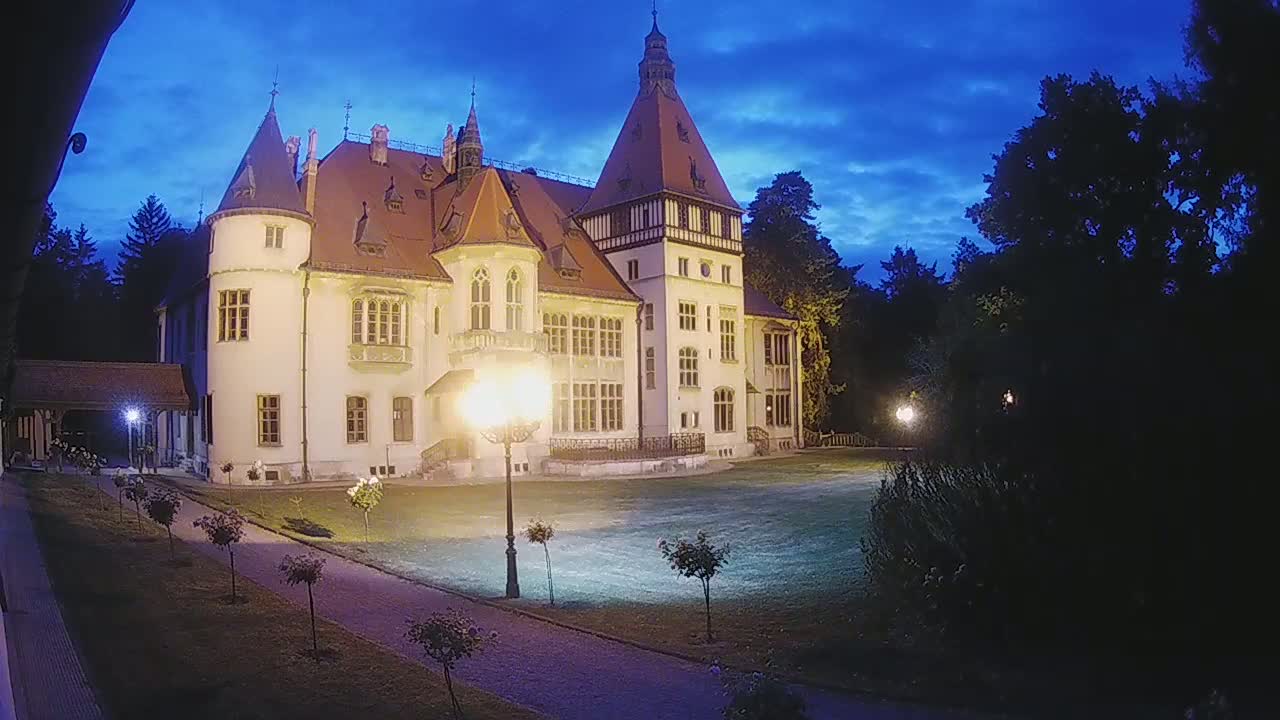 A large, light-colored castle with a red roof stands illuminated at twilight under a partly cloudy sky.
