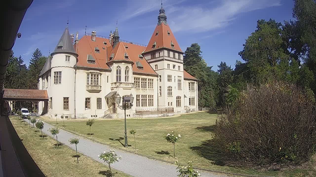 A large, light-colored castle with a red roof stands on a grassy lawn on a sunny day, surrounded by trees and shrubs.