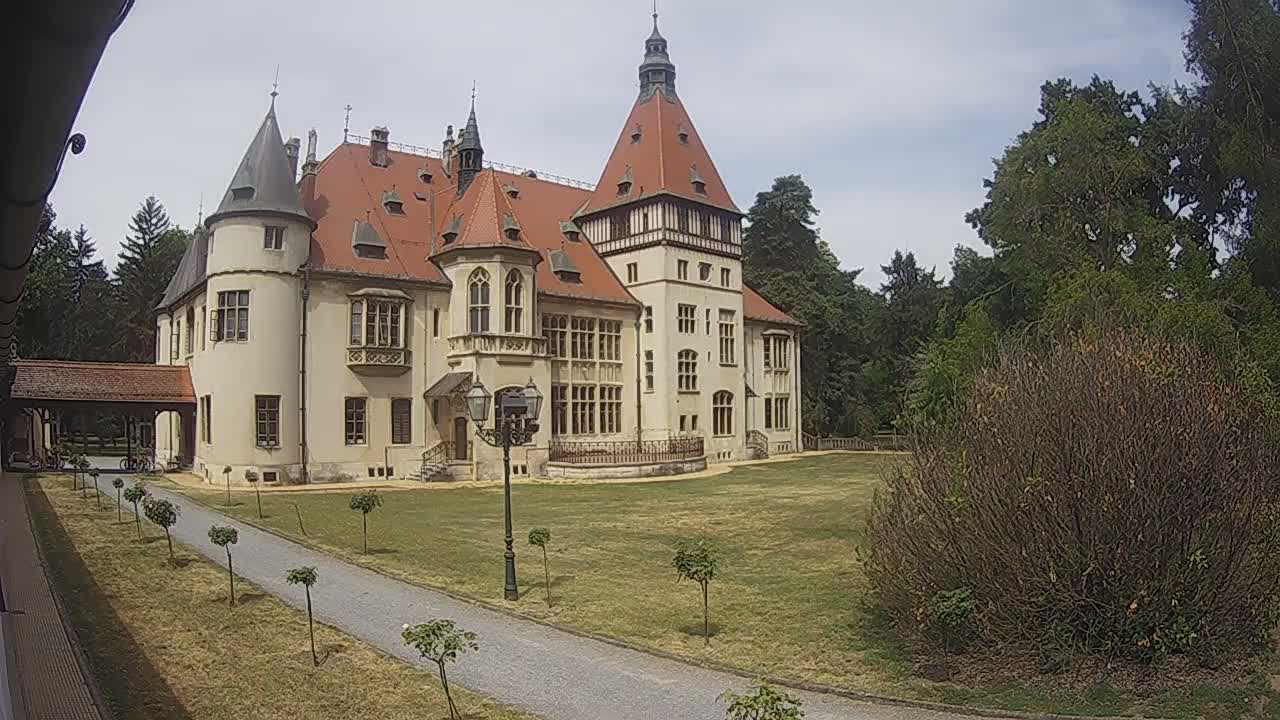 A large, light-colored castle with a red tile roof stands on a grassy lawn surrounded by trees under a partly cloudy sky.