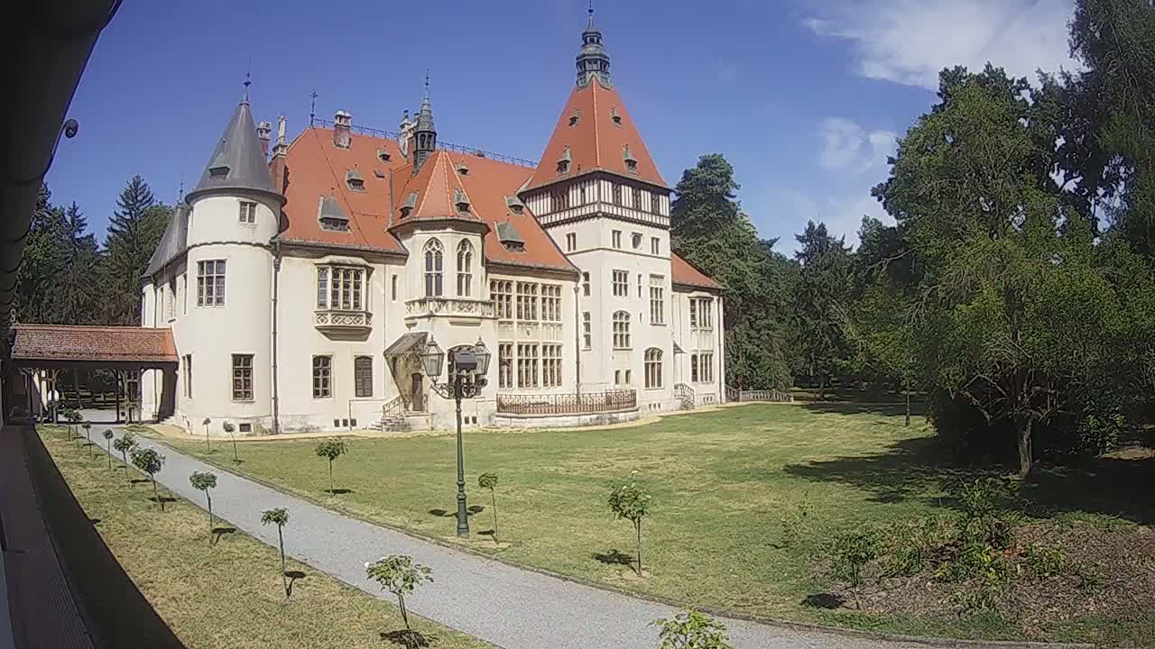 A large, light-colored castle with a red tile roof stands on a grassy lawn under a bright, sunny sky.