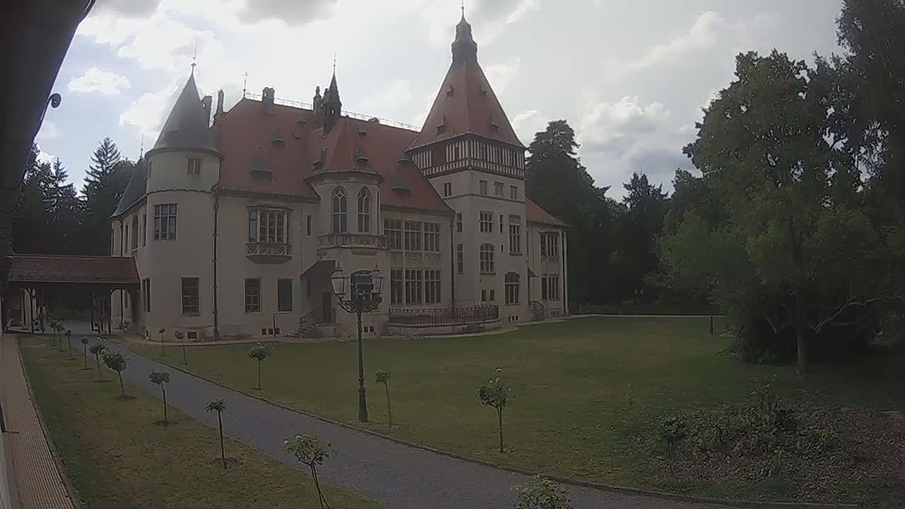A large, light-colored castle with a red tile roof stands on a grassy lawn under a bright, sunny sky.