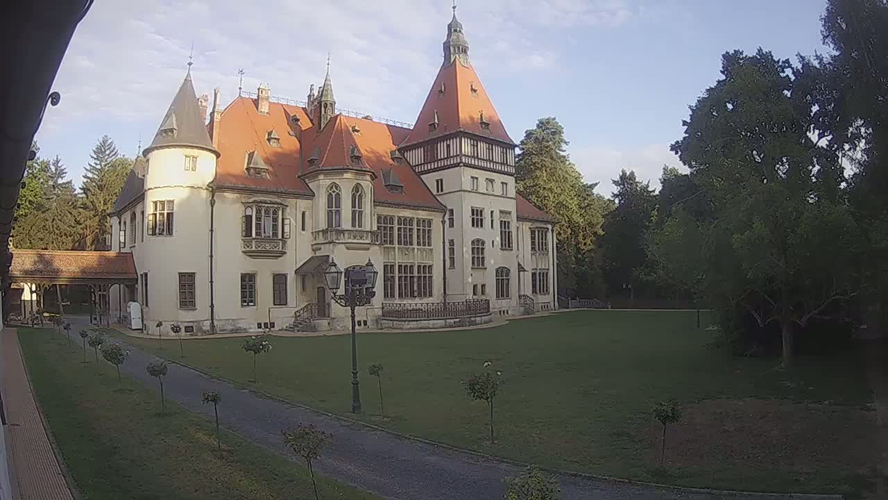 A large, light-colored castle with a red roof stands on a grassy lawn under a clear blue sky, and a person walks on a path nearby.